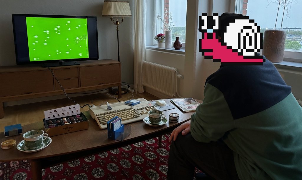 A man sitting in a sofa, with an Amiga 500 in front of him, watching Amiga demos while drinking coffee and eating chocolate.