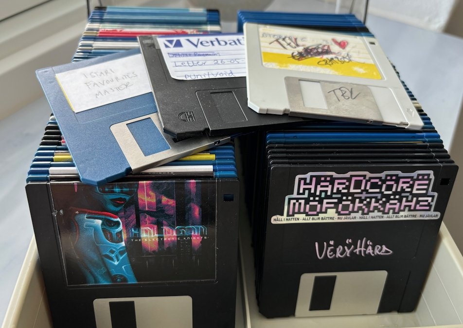 A close-up photo of a diskbox full of 3.5 inch floppy disks. On top of two orderly rows of disks in the box lies three floppies with messily-written disk labels.