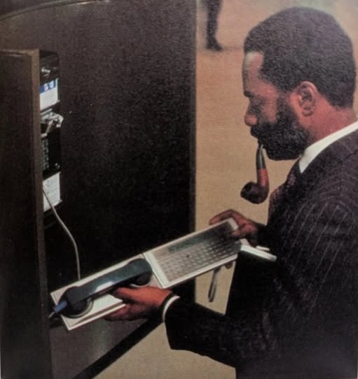 A dapper man is using an 1980s portable computer with a modem attachment at a public pay phone.